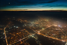 Aerial View From Airplane Window Of Buildings And Bright Illuminated Streets In City Residential Area At Night. Dark Urban Landscape At High Altitude