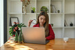 © saltdium - Photo of a young creative woman holding an eyeglasses and using a computer laptop at the wooden table surrounded by a coffee cup, stack of books and Christmas tree.