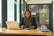 © saltdium - Photo of a young businesswoman holding a digital tablet while sitting at the wooden working desk surrounded by a computer laptop, house model and various office equipment.