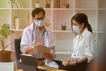 Photo of an older male general practitioner explaining a symptom to young patient woman at the table with a digital tablet.