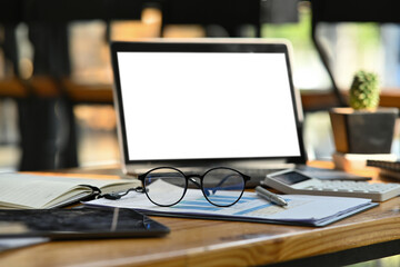 Poster - Photo of a comfortable working desk surrounded by a white blank screen computer laptop, eyeglasses, calculator, pen, notebook, digital tablet and document.