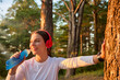 © Aleksandr - Close-up of a beautiful female fitness athlete in red wireless headphones who drinks water from a reusable soft sports bottle after a workout leaning against a tree