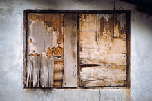 An Old Wooden Window Boarded Up With Cracked Plywood On A Concrete Wall. Old Aged Covered Cracked Wooden Panel Window Shutters. Easy Way To Repair Windows Of A Poor House. Plywood Collapse Background.