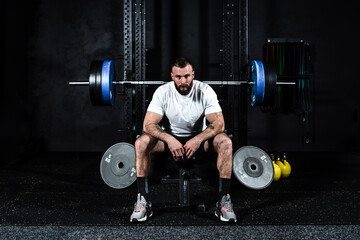 Young active strong sweaty muscular fit man with big muscles preparing for hardcore bench press workout cross training with heavy barbell weights in the gym. Male concentrated sitting on the bench.