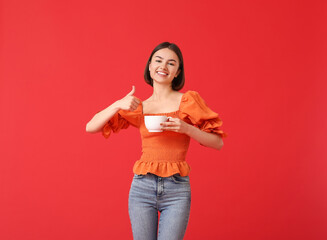 Wall Mural - Beautiful woman with cup of coffee showing thumb-up on color background