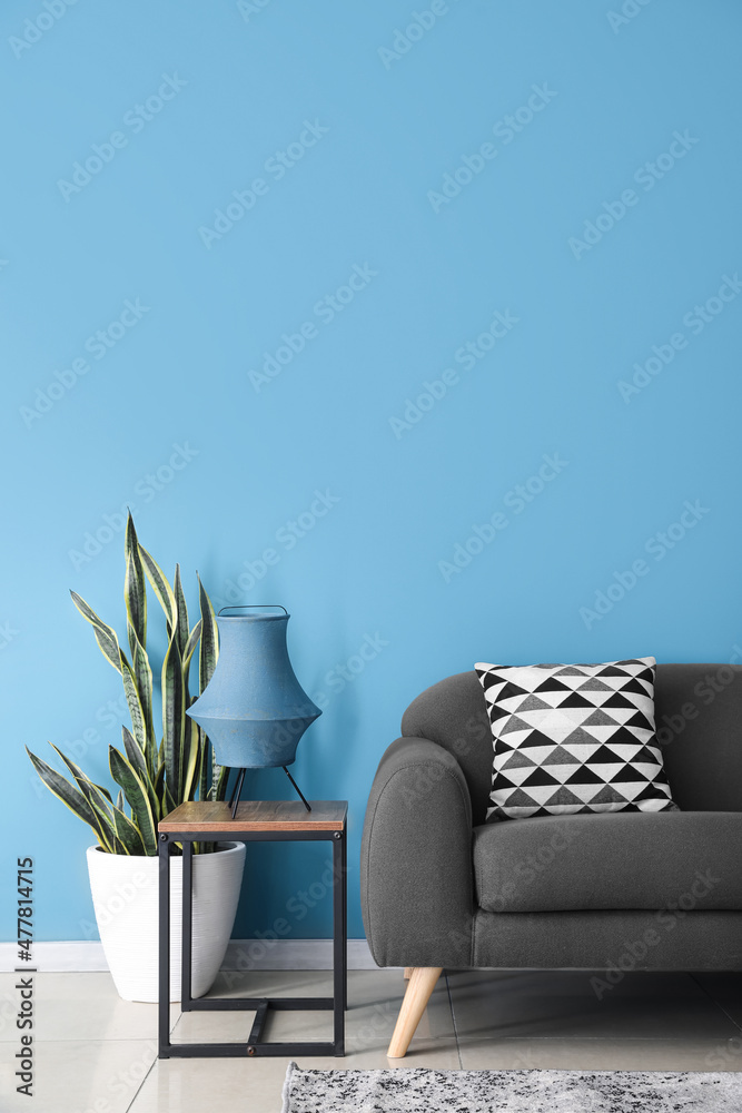 Stylish lamp on table, houseplant and sofa near blue wall