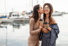Two Fun Women Are Kissing On The Cheek And Holding Cups Of Coffee In The Yacht Club.