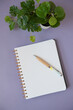 Cahier à spirale ouvert sur une page blanche avec un stylo et une plante verte sur un fond mauve pour trouver l'inspiration pour écrire