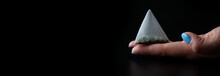 White Pyramid Tea Bag With Green Tea Lies In A Woman's Palm. Black Background. Macro. Web Banner. Selective Focus