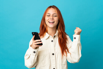 Wall Mural - Teenager Russian girl isolated on blue background using mobile phone and doing victory gesture