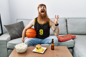 Poster - Caucasian man with long beard holding basketball ball cheering tv game crazy and mad shouting and yelling with aggressive expression and arms raised. frustration concept.