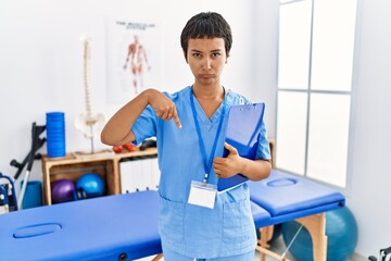 Wall Mural - Young hispanic woman with short hair working at pain recovery clinic pointing down looking sad and upset, indicating direction with fingers, unhappy and depressed.