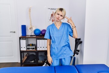 Wall Mural - Young physiotherapist woman working at pain recovery clinic shooting and killing oneself pointing hand and fingers to head like gun, suicide gesture.