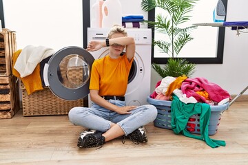 Sticker - Young blonde woman doing laundry sitting by washing machine covering eyes with arm, looking serious and sad. sightless, hiding and rejection concept