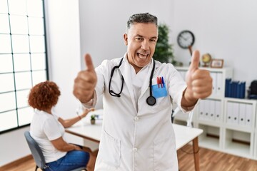 Poster - Middle age doctor man at the clinic with a patient approving doing positive gesture with hand, thumbs up smiling and happy for success. winner gesture.