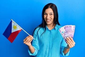 Poster - Young latin woman holding philippines flag and pesos banknotes clueless and confused expression. doubt concept.