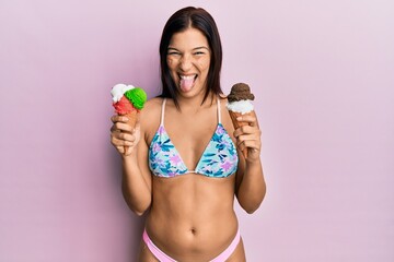 Sticker - Young latin woman wearing bikini holding ice cream sticking tongue out happy with funny expression.