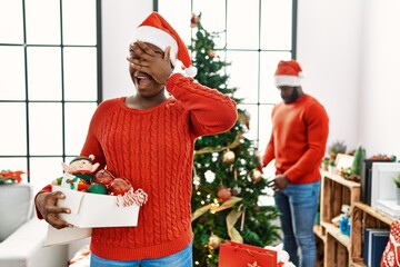 Canvas Print - Young african american couple standing by christmas tree peeking in shock covering face and eyes with hand, looking through fingers with embarrassed expression.