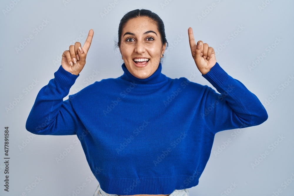 Young hispanic woman standing over isolated background smiling amazed and surprised and pointing up with fingers and raised arms.
