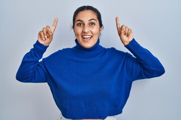 Poster - Young hispanic woman standing over isolated background smiling amazed and surprised and pointing up with fingers and raised arms.