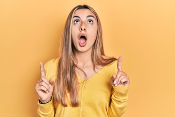 Wall Mural - Beautiful hispanic woman wearing casual yellow sweater amazed and surprised looking up and pointing with fingers and raised arms.