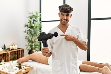 Wall Mural - Young hispanic man holding therapy massage gun at wellness center smiling happy pointing with hand and finger