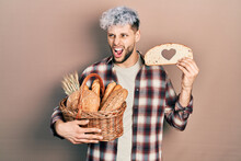 Young Hispanic Man With Modern Dyed Hair Holding Wicker Basket With Bread And Loaf With Heart Symbol Angry And Mad Screaming Frustrated And Furious, Shouting With Anger. Rage And Aggressive Concept.