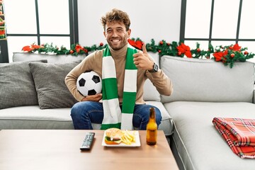 Sticker - Young hispanic man football hooligan holding ball at home smiling happy and positive, thumb up doing excellent and approval sign