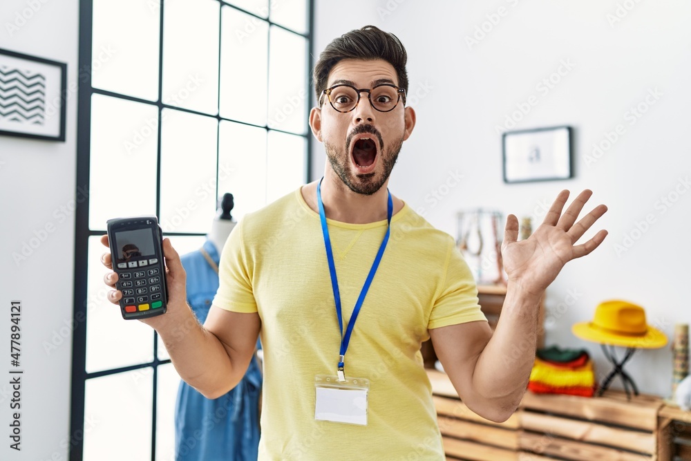 Young man with beard working at retail boutique holding dataphone scared and amazed with open mouth for surprise, disbelief face