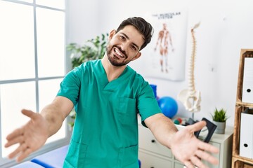 Poster - Young man with beard working at pain recovery clinic looking at the camera smiling with open arms for hug. cheerful expression embracing happiness.