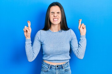 Young brunette teenager wearing casual sweater gesturing finger crossed smiling with hope and eyes closed. luck and superstitious concept.