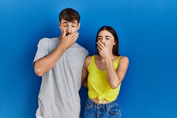 Poster - Young hispanic couple standing together over blue background bored yawning tired covering mouth with hand. restless and sleepiness.