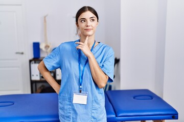 Canvas Print - Young hispanic woman wearing physiotherapist uniform standing at clinic thinking concentrated about doubt with finger on chin and looking up wondering