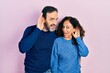 Middle age couple of hispanic woman and man hugging and standing together smiling with hand over ear listening an hearing to rumor or gossip. deafness concept.