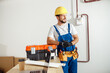 © Kostiantyn - Smiling electrician, handyman in uniform, hard hat and protective gloves looking aside while posing indoors, leaning on a toolbox