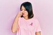 Young hispanic woman wearing pink cancer ribbon on t shirt smelling something stinky and disgusting, intolerable smell, holding breath with fingers on nose. bad smell