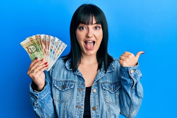 Sticker - Young hispanic woman holding peruvian sol banknotes pointing thumb up to the side smiling happy with open mouth