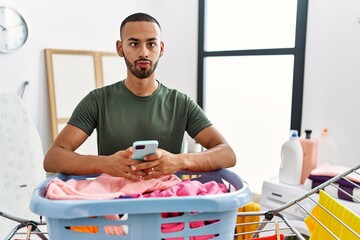 Poster - African american man doing laundry using smartphone making fish face with mouth and squinting eyes, crazy and comical.