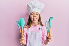 Beautiful Brunette Little Girl Wearing Professional Cook Apron Holding Cooking Tools Sticking Tongue Out Happy With Funny Expression.