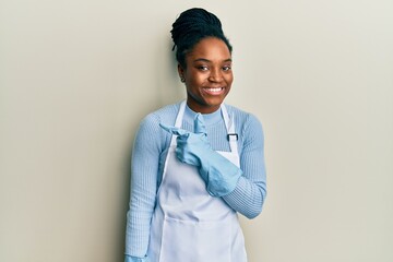 Wall Mural - African american woman with braided hair wearing cleaner apron and gloves cheerful with a smile of face pointing with hand and finger up to the side with happy and natural expression on face