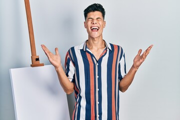 Canvas Print - Young hispanic man standing close to empty canvas celebrating mad and crazy for success with arms raised and closed eyes screaming excited. winner concept