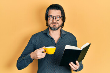 Wall Mural - Middle age caucasian man reading book drinking cup of tea puffing cheeks with funny face. mouth inflated with air, catching air.