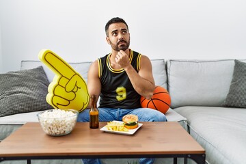 Wall Mural - Young hispanic man with beard holding basketball ball cheering tv game serious face thinking about question with hand on chin, thoughtful about confusing idea