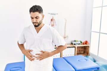 Wall Mural - Young handsome man with beard working at pain recovery clinic with hand on stomach because indigestion, painful illness feeling unwell. ache concept.