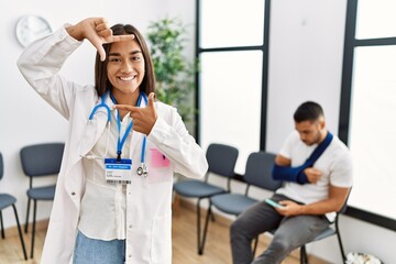 Canvas Print - Young asian doctor woman at waiting room with a man with a broken arm smiling making frame with hands and fingers with happy face. creativity and photography concept.