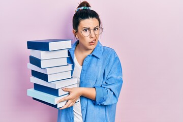 Poster - Young hispanic woman wearing glasses and holding books clueless and confused expression. doubt concept.