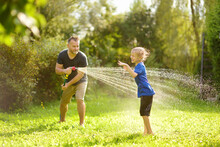 Funny Little Boy With His Father Playing With Garden Hose In Sunny Backyard. Preschooler Child Having Fun With Spray Of Water. Summer Outdoors Activity For Kids.