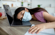 Unhappy Indian Teen Girl In Face Mask Lying On Laptop Keyboard, Feeling Bored Of Online Studies During Covid Quarantine