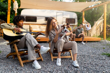 Young Black Guy Playing Guitar And Singing, Caucasian Lady Holding Dog Near RV, Camping With Diverse Friends In Summer