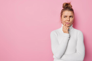 Wall Mural - Horizontal shot of discontent sullen young woman looks thoughtfully away purses lips wears casual white turtleneck isolated over pink background with copy space for your promotional content.